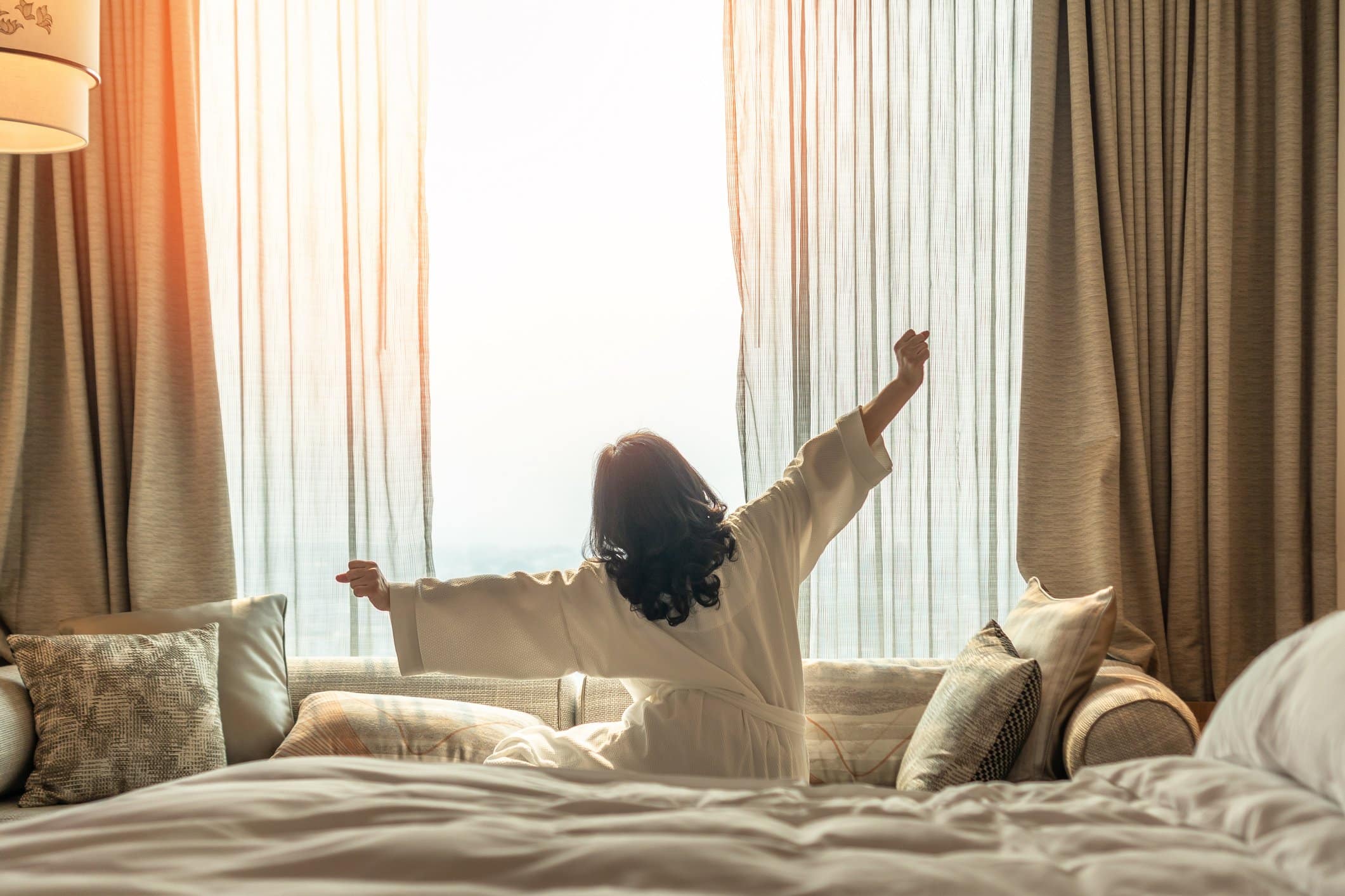 Woman stretching with arms outstretched while sitting on bed in front of large window with curtains.