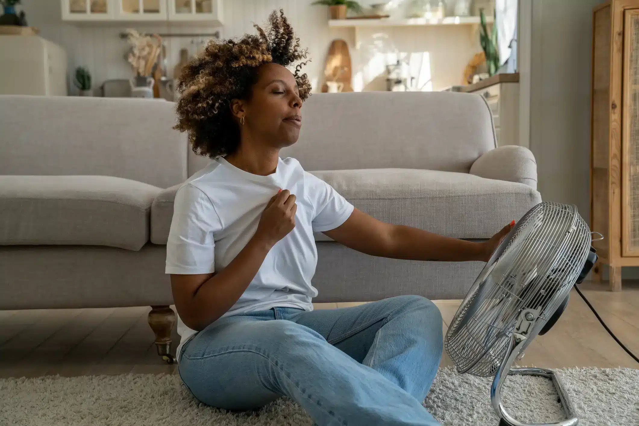A woman with curly hair sitting on the floor with her eyes closed, holding her chest with one hand, in front of an electric fan.