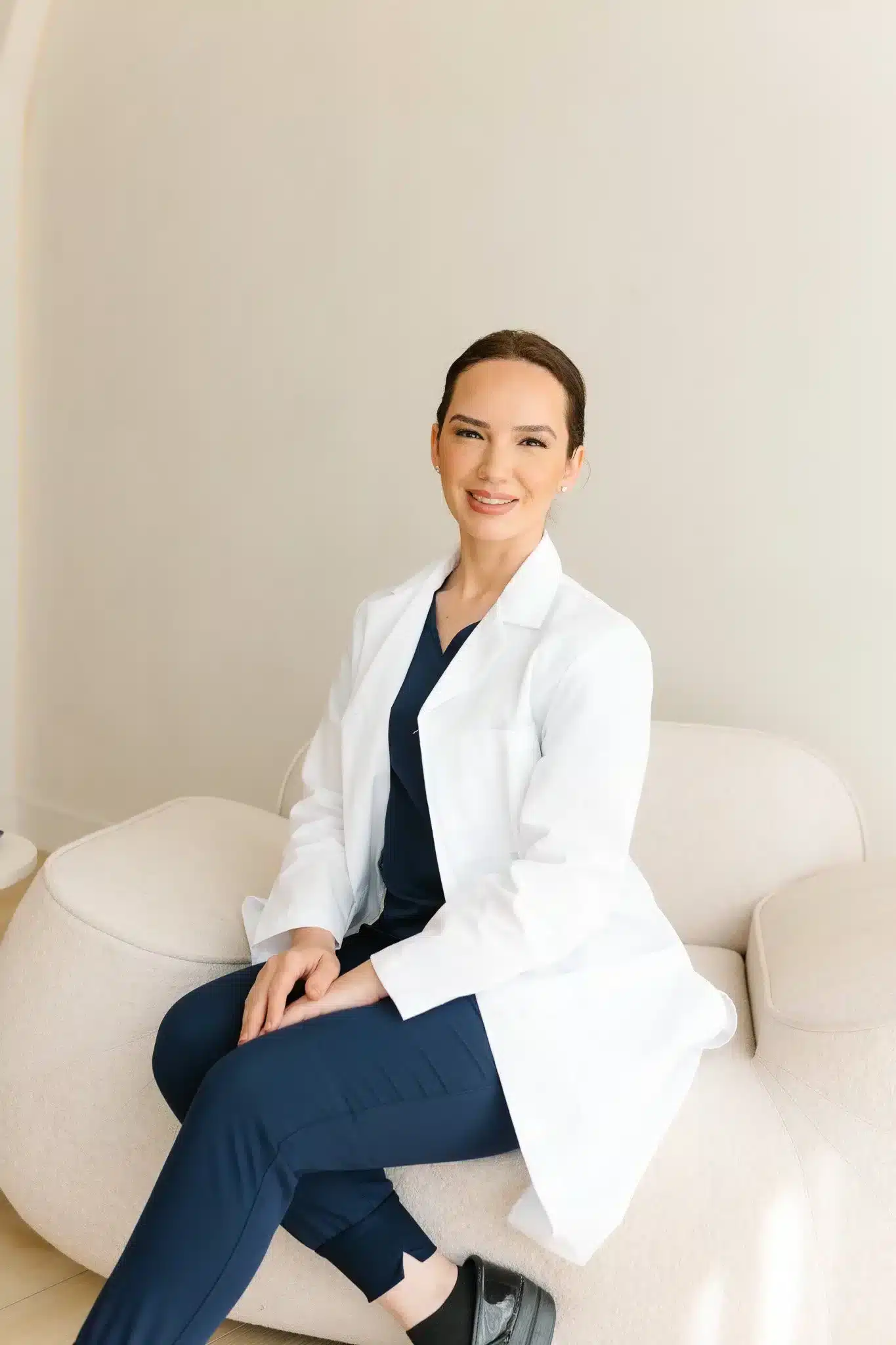 A woman in a white coat sitting on a cream-colored sofa, smiling at the camera, with a neutral background.